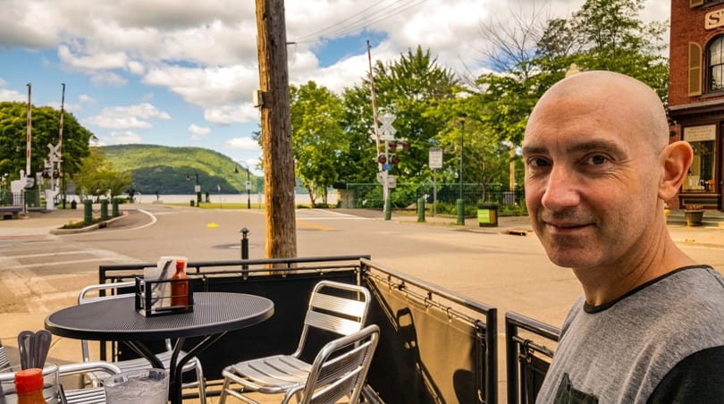 Man on a restaurant patio with a scenic view of a river, mountains, and a road.